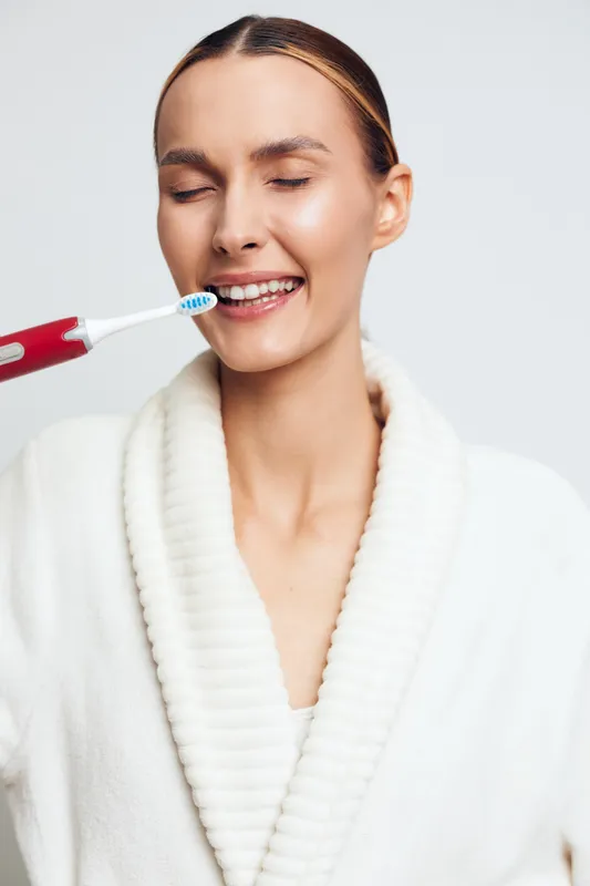 A young woman in a white bathrobe smiles with closed eyes while holding an electric toothbrush near her mouth. The image represents dental care and morning routine concepts.