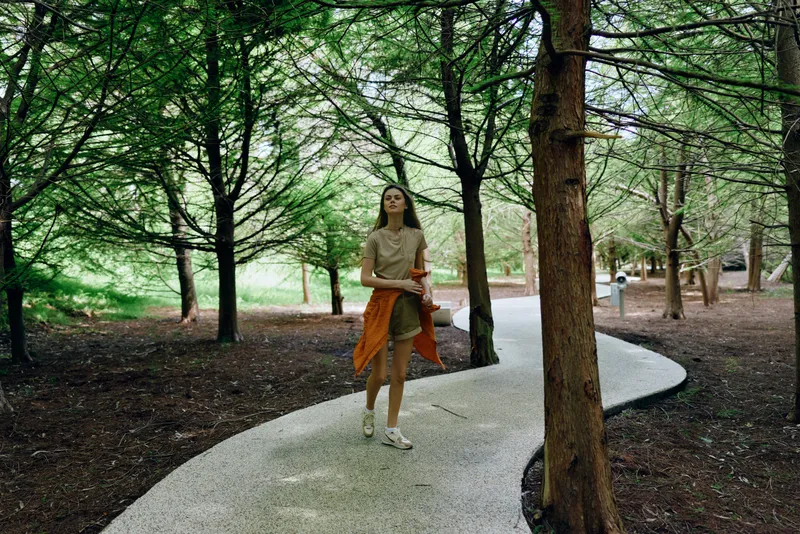 woman walking in a park on a winding path among trees and nature, wearing a casual dress and sneakers with an orange jacket tied at waist, serene outdoor lifestyle portrait