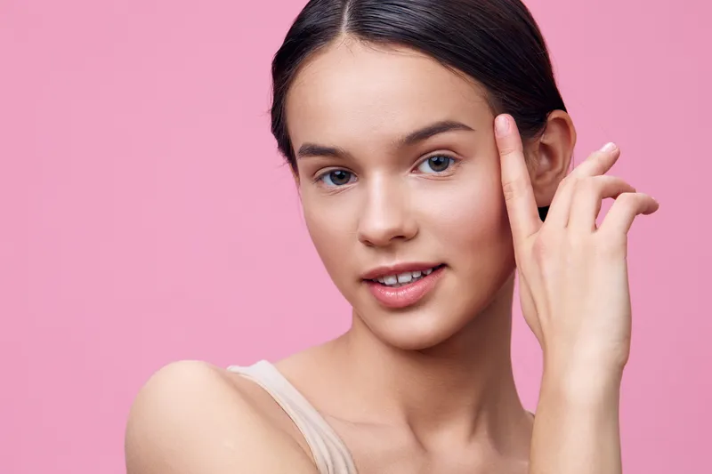 Young woman smiling confidently against a pink background, showcasing natural beauty and effortless style Soft makeup enhances her radiant skin tone