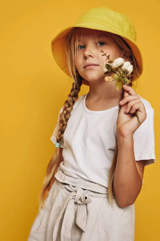 Girl with long braided hair wears a yellow hat and white t-shirt holding a small bouquet on a yellow background. Studio portrait of thoughtful child.