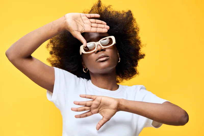 Young Black woman with curly hair wearing stylish sunglasses poses playfully against a vibrant yellow background, exuding confidence and joy