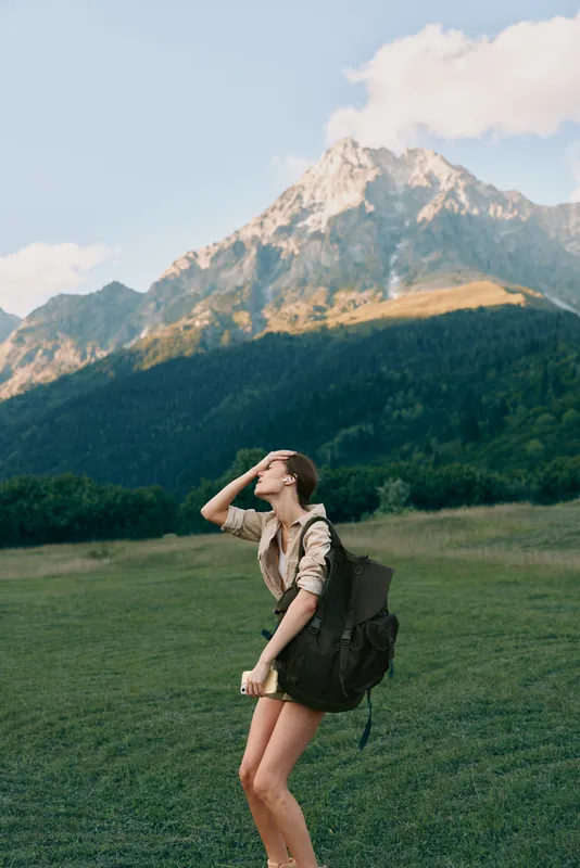 Woman traveler with a backpack stands in a grassy meadow, gazing towards distant mountains, capturing a lifestyle moment of hiking outdoors amid nature