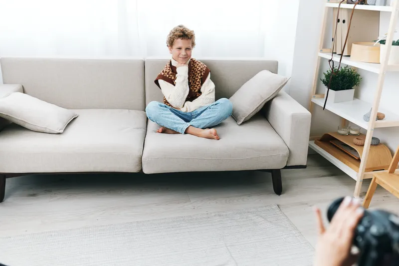 Young boy sits cross legged on a light grey sofa, posing for a cheerful portrait in a bright living room with soft cushions and clean decor.