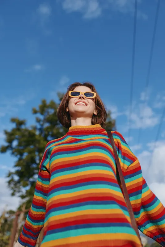 Woman in a colorful striped sweater wearing yellow sunglasses, smiling outdoors under a bright blue sky; young casual outfit with shoulder bag, joyful travel lifestyle portrait.