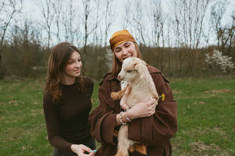 Two women share a warm moment in a grassy park, one cradles a fluffy goat kid, wearing earthy tones, casual coats, conveying friendly, outdoor lifestyle and tender animal connection. They smile