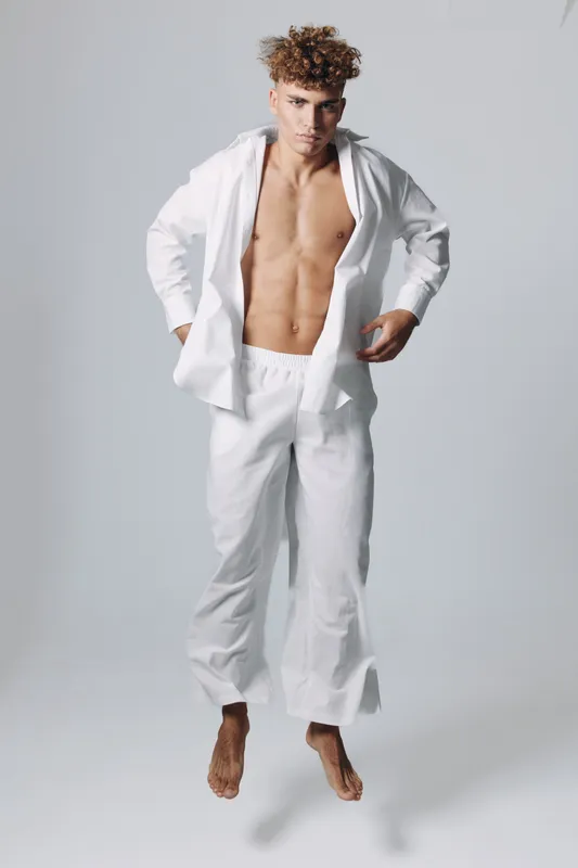 Young man with curly hair wearing white loose pants and open shirt jumping barefoot against plain background. Athletic body, casual style and dynamic pose in studio lighting.