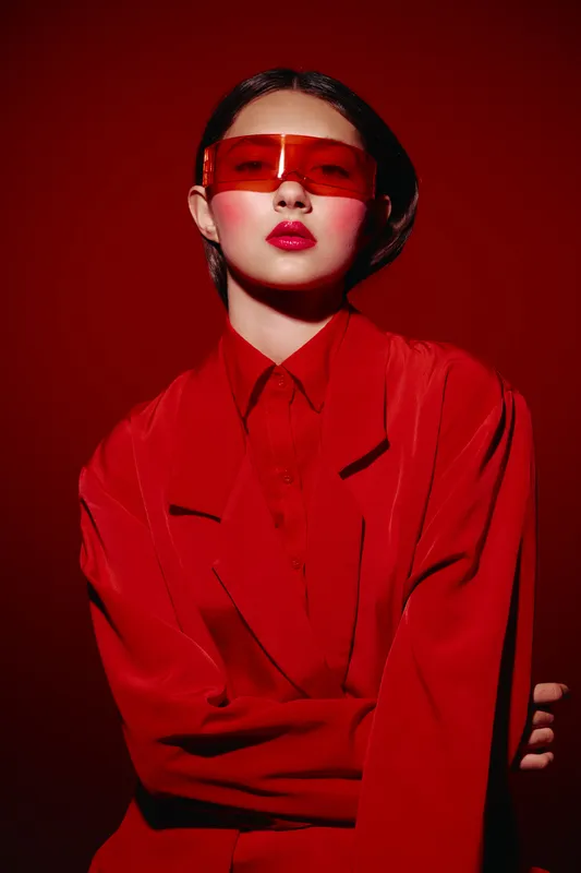 Young woman wearing a bold red outfit and translucent red sunglasses looks confidently at the camera with styled hair. Studio shot with dramatic lighting. Beauty creative fashion concept.