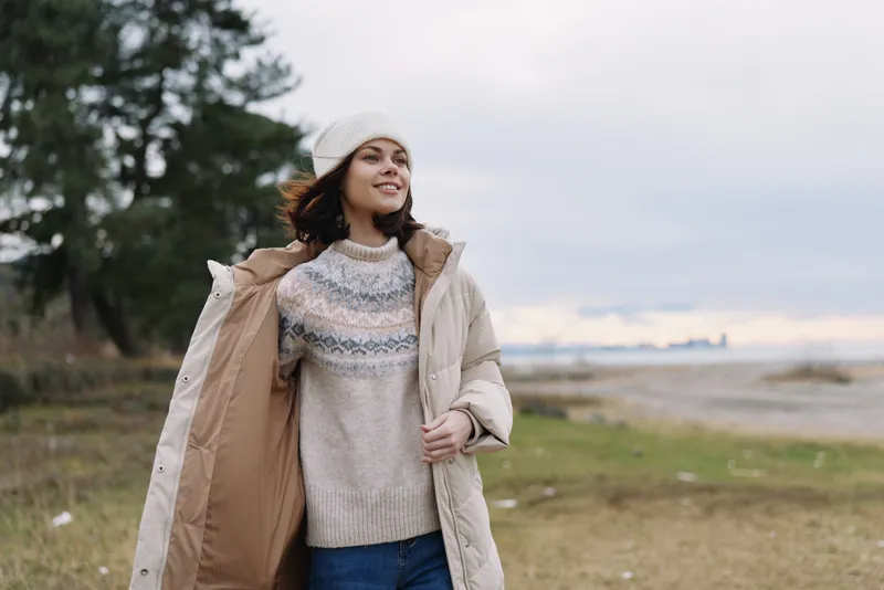 Woman in a knit sweater and beige coat smiles outdoors along a windy coast, wearing a casual jacket and jeans by the shoreline.