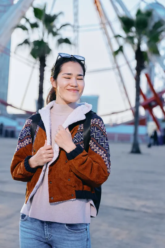 Smiling woman wearing a textured brown jacket with shearling lining, sunglasses atop head, standing outdoors on a sunny street with palm trees, casual urban fashion, relaxed pose, candid lifestyle