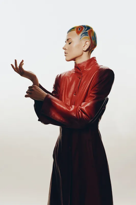 Young woman with artistic colorful hair painting wearing a long red leather coat standing elegantly against a light background in a minimalist studio setting.