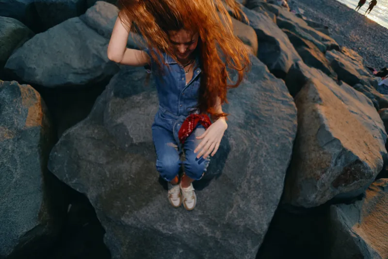 redhead woman jumping on large rocky shore wearing casual denim jumpsuit and sneakers during sunny outdoor activity near water