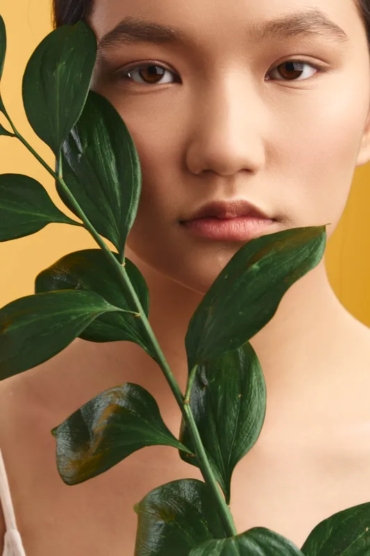 asian woman with delicate features is seen through glossy green leaves, creating a serene portrait with natural texture, soft light, and a warm yellow background that enhances mood and focus.