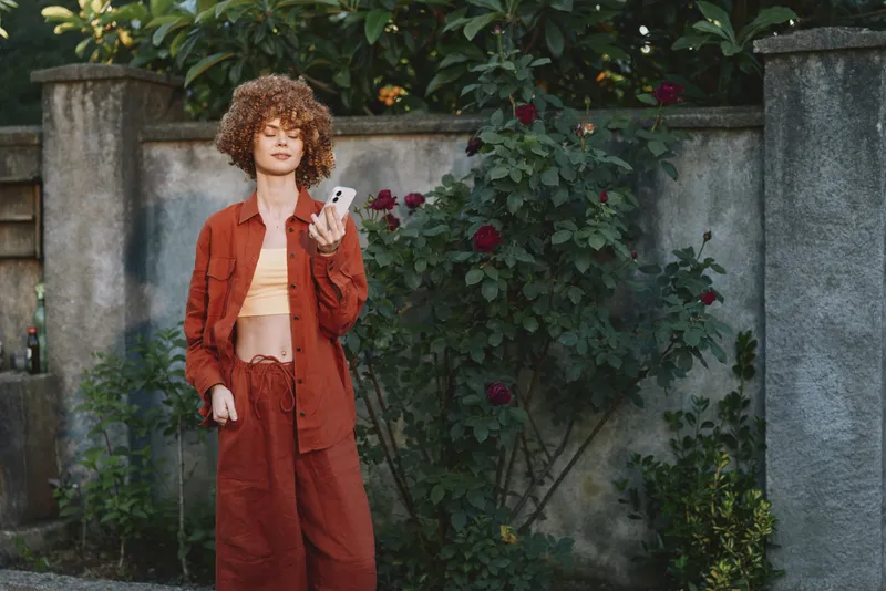 Curly haired woman in an orange suit standing outdoors with a phone, portraying contemporary lifestyle, confidence, and self expression in daylight