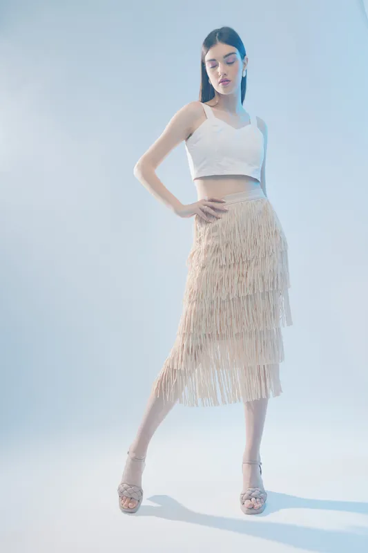Young woman in a white crop top and beige fringe skirt poses confidently on a light background. Concept of woman beauty fashion, elegance, and modern style.