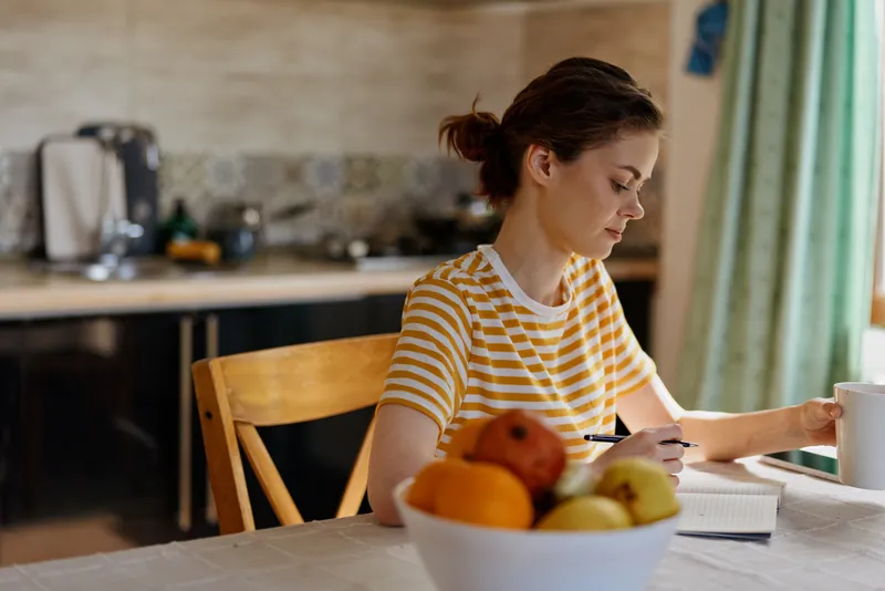 Writer in a striped yellow shirt sits at a kitchen table writing in a notebook beside a bowl of fruit, a warm cozy home scene.