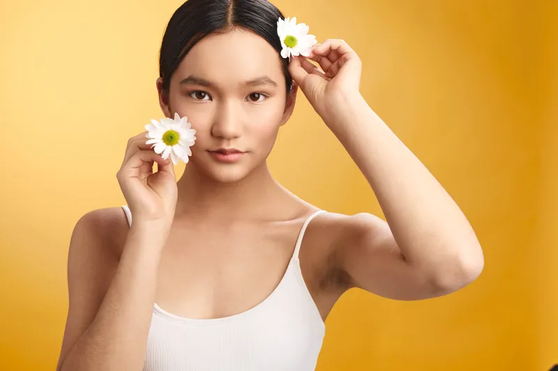 Asian woman holds daisies against a warm yellow backdrop, creating a fresh portrait with soft lighting. Her calm expression radiates gentle happiness, evoking natural beauty and serene optimism.
