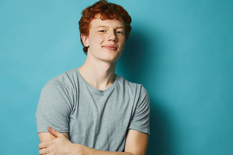 Young man with red hair and freckles smiling confidently with arms crossed wearing a gray t shirt on blue background. Casual portrait of happy male with relaxed posture.