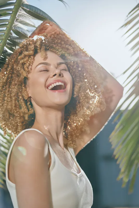 smiling woman with curly hair enjoying sunlight outdoors near palm leaves, wearing white sleeveless top, radiant joyful expression in bright natural setting