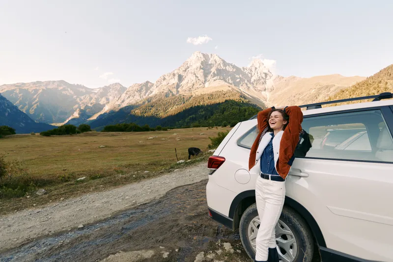 Woman leans against car at roadside with mountain view, nature travel scene and relaxed outdoors vibe, smiling in jacket under sunlight during scenic road trip across open landscape.