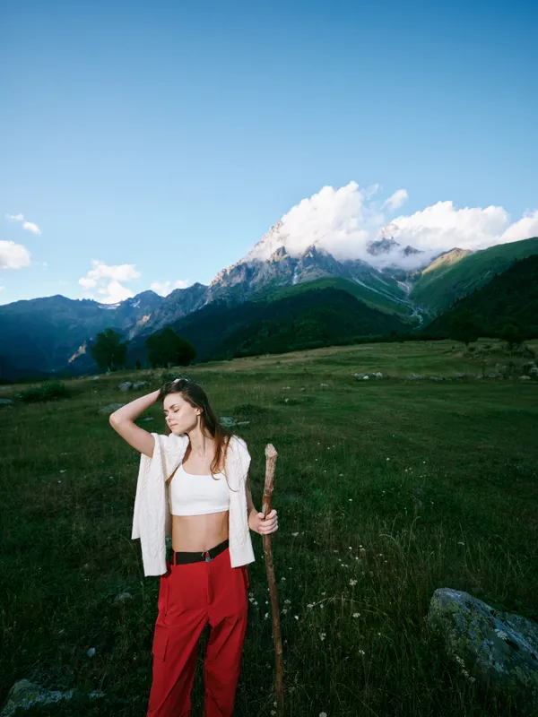 Woman standing in a grassy meadow with a wooden staff, white top and red pants, mountains and blue sky behind for an outdoor adventure scene