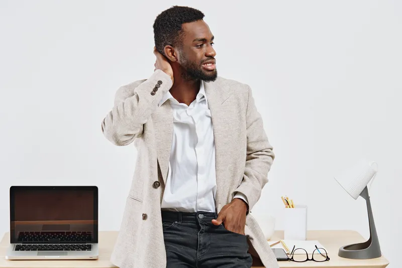 Confident young man with dark skin and curly hair in smart casual clothing standing at modern office desk, holding neck and smiling while looking sideways. Workplace setting, professional lifestyle co