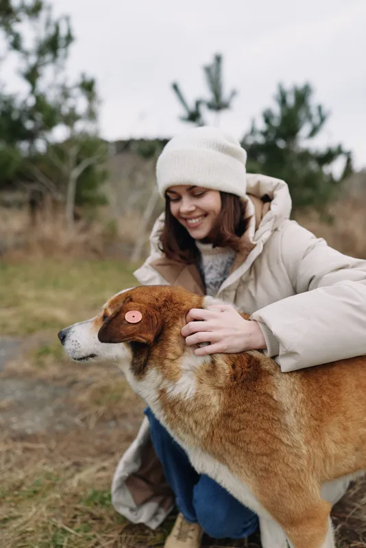 Woman in a cozy coat and beanie gently pets a dog outdoors, sharing affection in a natural field on a chilly day, capturing a tender moment of companionship