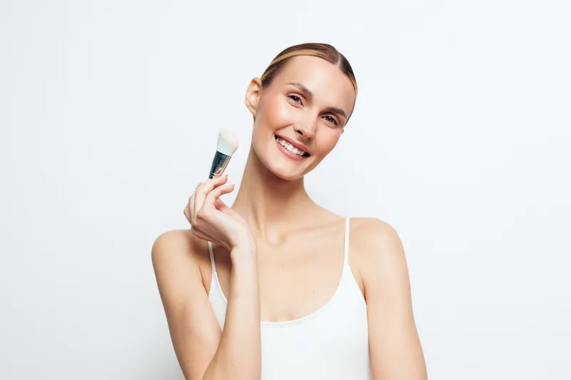 Young woman with blonde hair smiles while holding a makeup brush near her face against a white background in a bright and clean studio setting.
