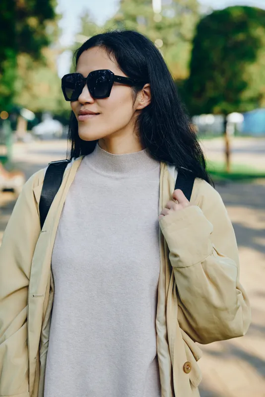 Woman in beige coat and sweater wearing sunglasses in a sunny park, casual street style, relaxed mood, daytime outdoor fashion, natural light, soft shadows, candid pose, minimal background, urban