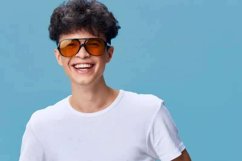 Happy man wearing orange sunglasses and a white t-shirt smiling broadly against a blue background, expressing joy and youthful energy with curly hair.