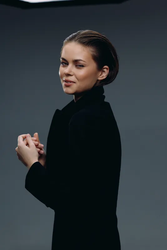Portrait of confident young woman wearing black turtleneck with natural makeup and slicked-back hair against dark background.