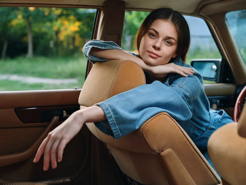 A beautiful young woman in a denim jacket resting in a vintage car, showcasing a serene expression against a lush green background in soft daylight.