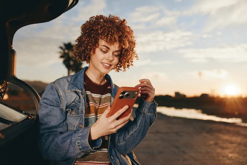 Smiling woman with curly hair holding a smartphone beside a car in a denim jacket at sunset. Outdoor travel lifestyle with warm golden sunlight and soft focus for a relaxed mood.