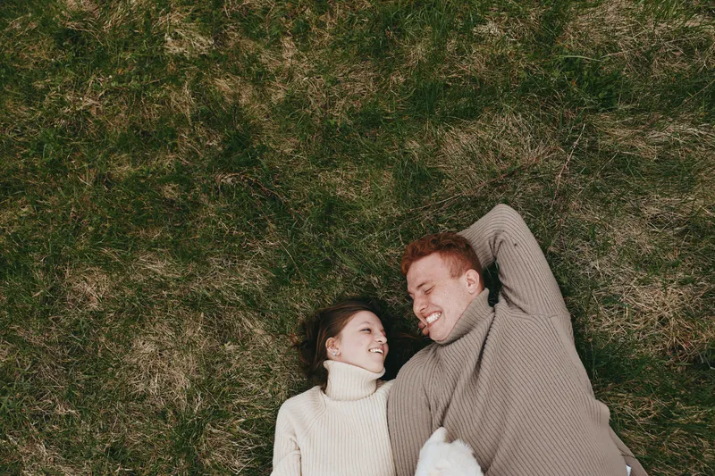 Young couple in cozy sweaters lies on green grass looking at each other with smiles enjoying a relaxed lifestyle moment outdoors in autumn season.