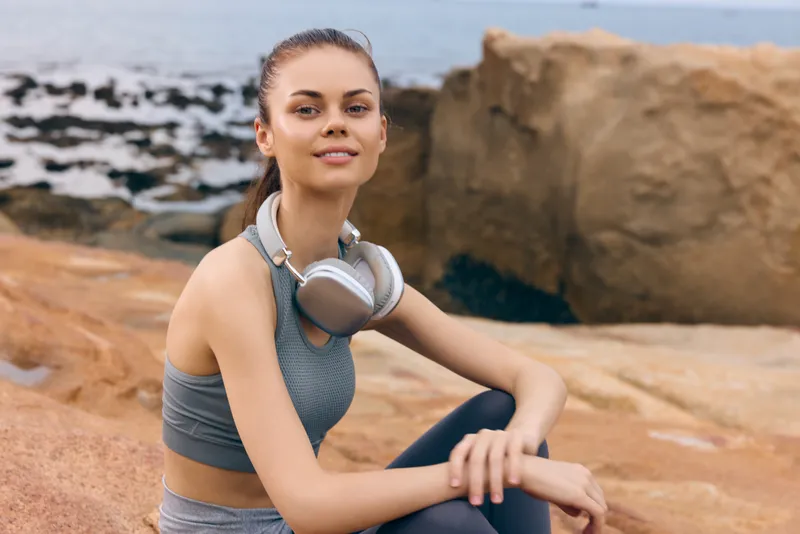 Young athletic woman enjoying outdoors, wearing headphones, sitting on the rocks near the sea, promoting a healthy lifestyle and physical fitness