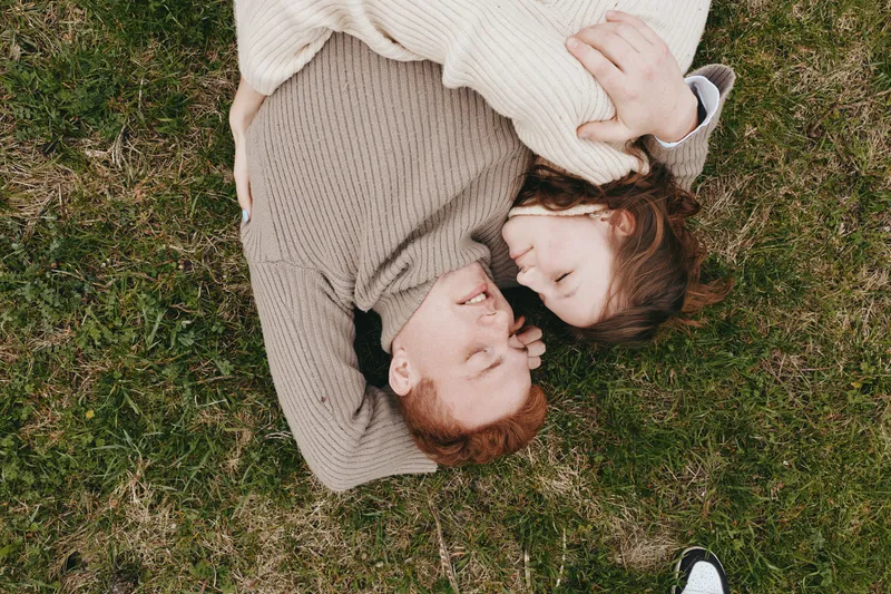 Relaxed couple lying on grass outdoors, smiling and embracing, cozy sweaters, warm light, intimate moment in a park setting, casual lifestyle scene highlighting connection and comfort.
