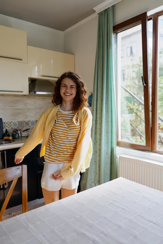 Woman in striped shirt and yellow cardigan stands in a bright home kitchen, smiling warmly, enjoying a casual morning routine, relaxed posture, daylight flooding the room and cozy atmosphere