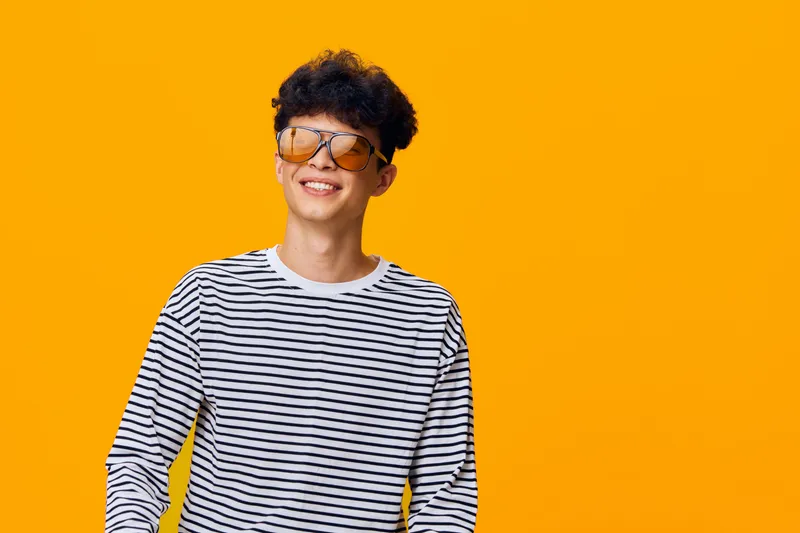 Young man smiling confidently wearing striped shirt and stylish sunglasses against bright yellow background, showcasing casual modern fashion and positive mood.