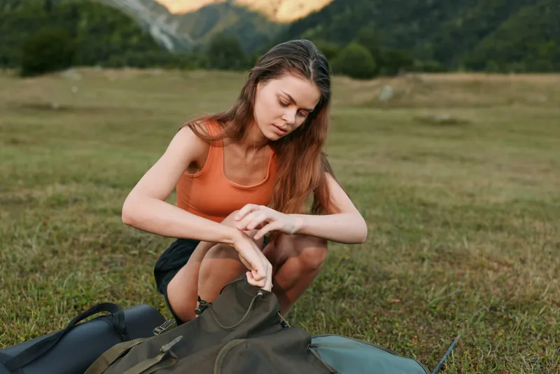 A young woman outdoors in a grassy field packs a backpack, preparing gear for a hike. She wears an orange top and shorts, focused and calm amid natural surroundings.