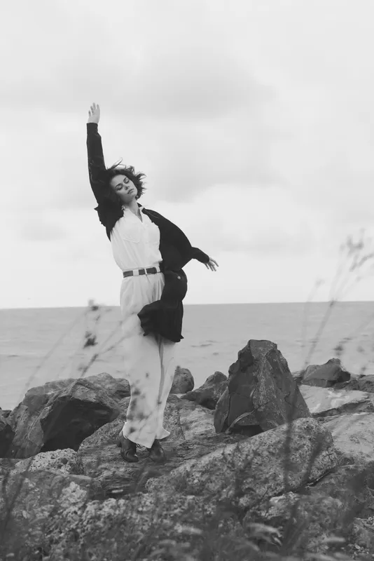 Dance, woman on a rocky coastline by the sea, flowing dress, wind catching fabric, graceful pose with raised arm, monochrome mood, sea breeze, outdoor motion, artistic expression and freedom in nature