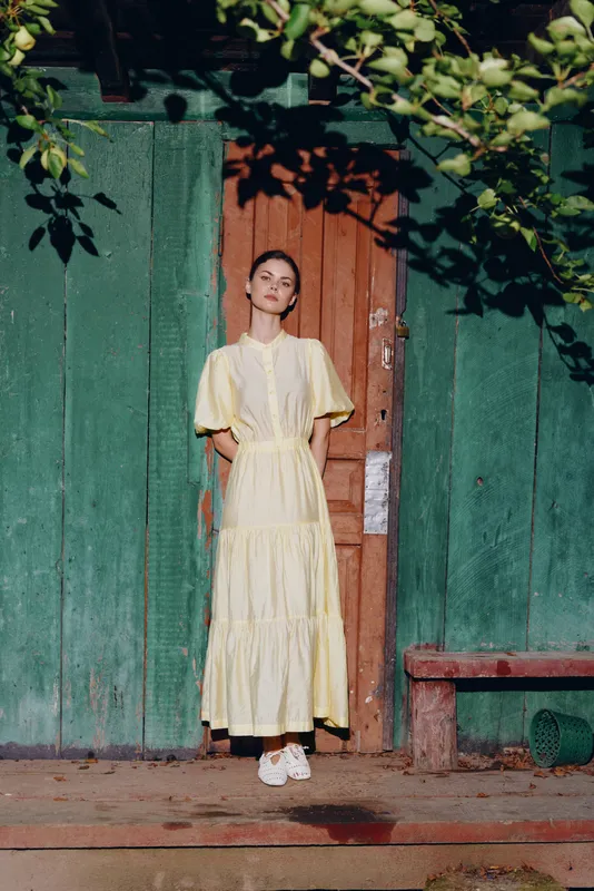 Elegant woman in a flowing yellow dress stands before a green wooden doorway outdoors, a calm garden ambiance and soft sunlight highlighting her pose.