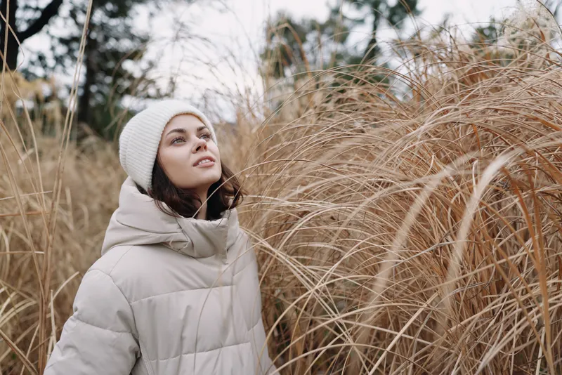 Woman in a warm coat and knit hat stands among tall grasses in a serene outdoor field, capturing a quiet reflective moment in soft natural light.