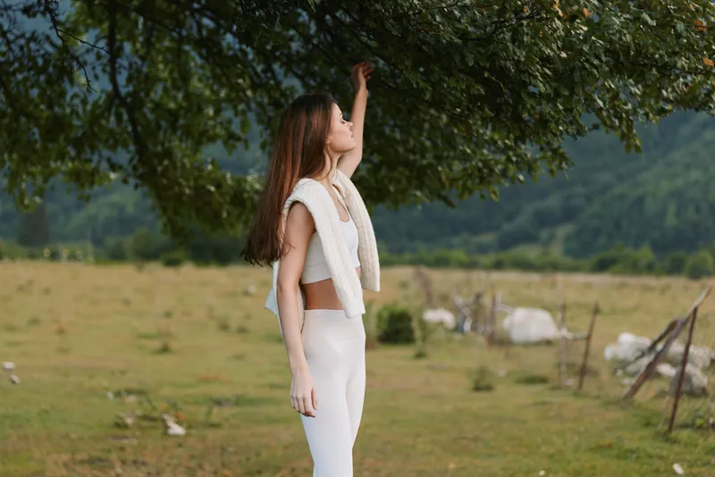 Young woman in white outfit standing in a green field reaching up to tree branches. Outdoor lifestyle scene with nature and wellness concept in soft natural light.