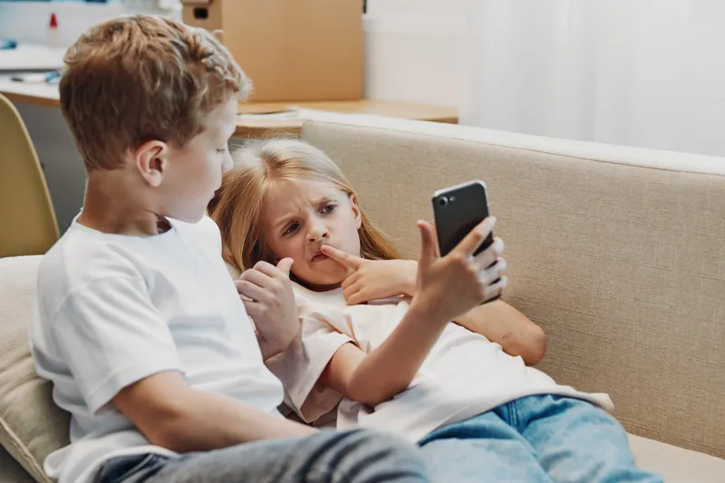 Two children relax on a beige sofa, a boy and a girl using a smartphone together in a cozy living room with natural light. Concept of childhood, technology, and casual moments.