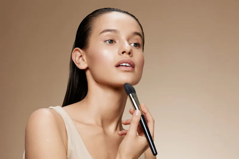 Young woman applying makeup with a brush on a neutral background, showcasing flawless skin and a natural beauty concept, ideal for beauty advertising and skincare