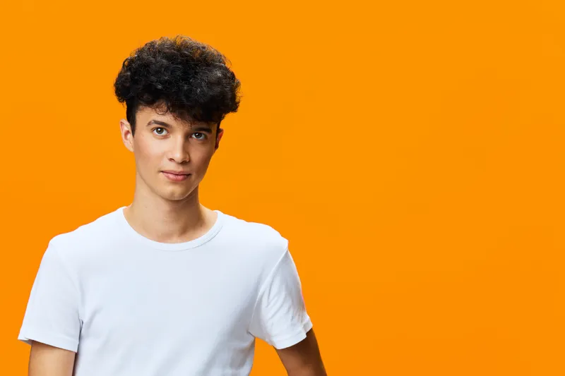 young man with curly hair wearing casual white t-shirt stands against vibrant orange background, looking confidently at camera with relaxed expression