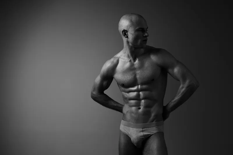 Black and white image of a muscular man posing confidently, showcasing strength and fitness in a minimalist studio background.