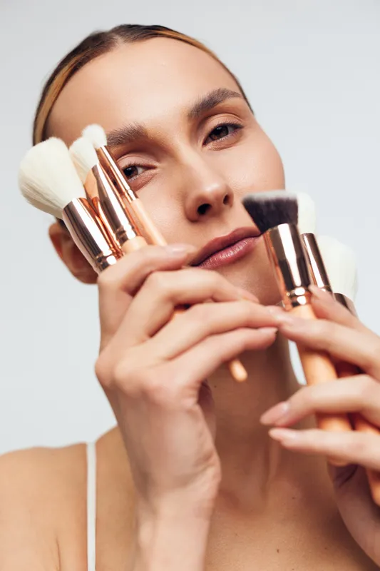 Young woman with makeup brushes near her face, soft studio light, focused expression, beauty tools in hand, clean background, natural tones, intimate closeup of makeup routine