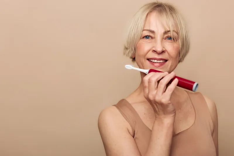 Smiling mature woman with short blonde hair holding electric toothbrush near face on beige background. Concept for dental care, oral hygiene, healthy lifestyle, and self-confidence at home.