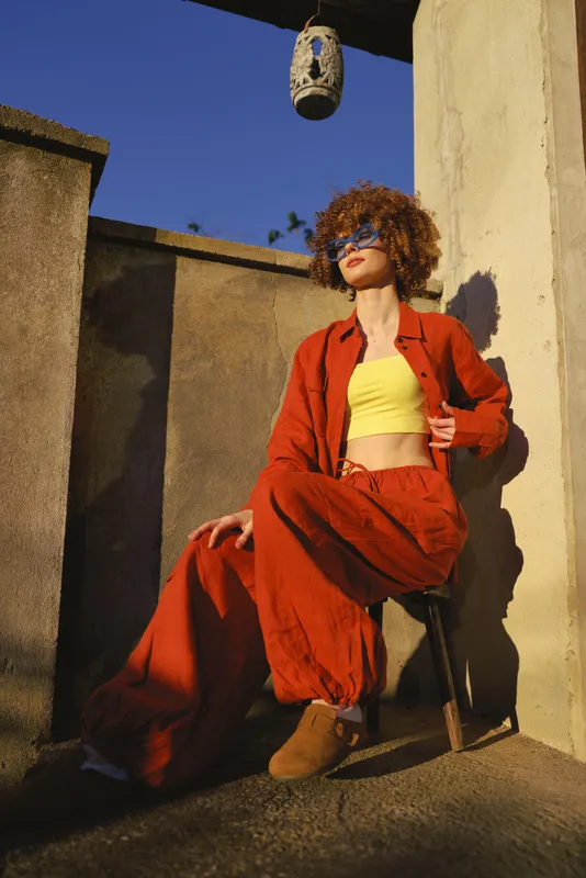 Vibrant woman outdoor fashion in bold red jumpsuit with yellow crop top, sunglasses, relaxed pose, sunlit atmosphere against urban wall backdrop