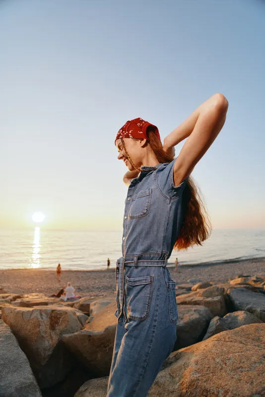 lifestyle woman in modern boho-western denim street style stretching by the rocky beach during warm golden hour with film color tones and relaxed mood
